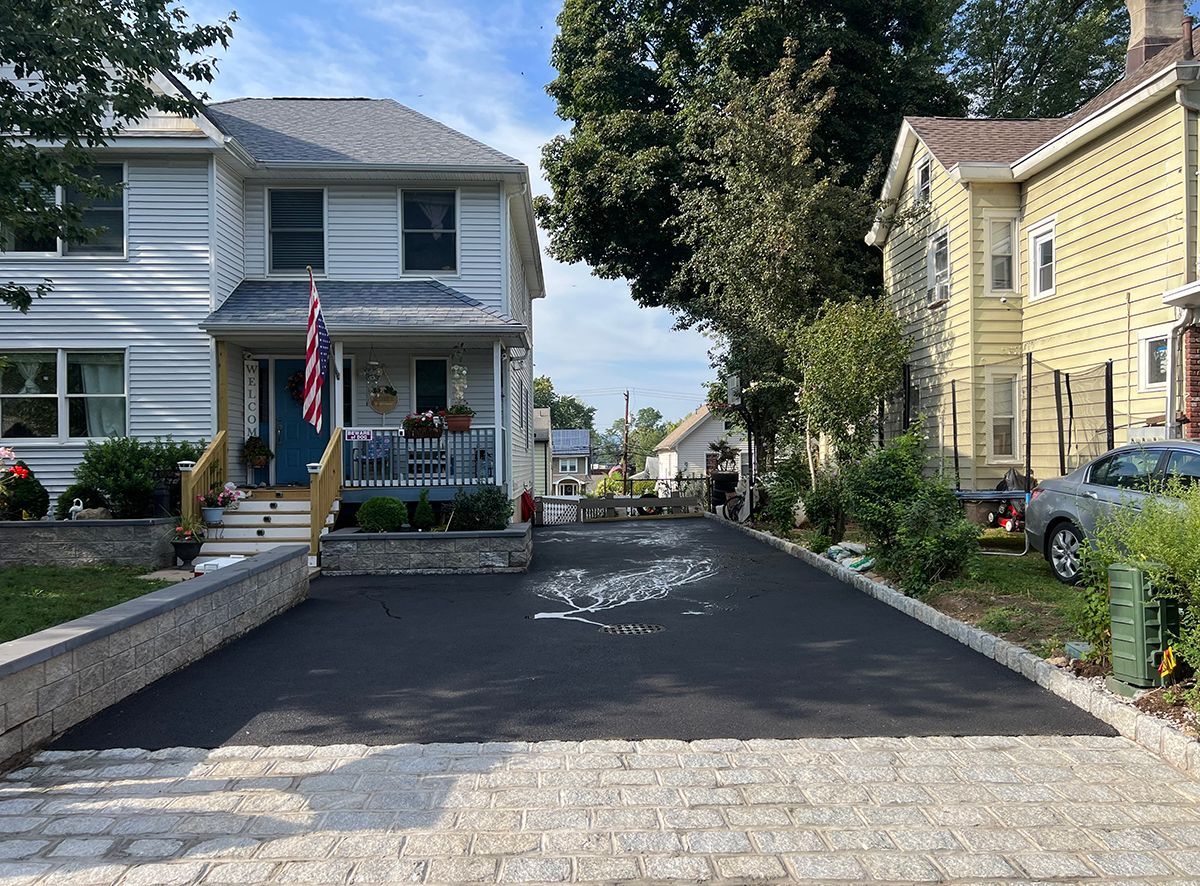 Paved driveway between two houses. One is white with a porch, the other yellow. Trees and cars are visible.