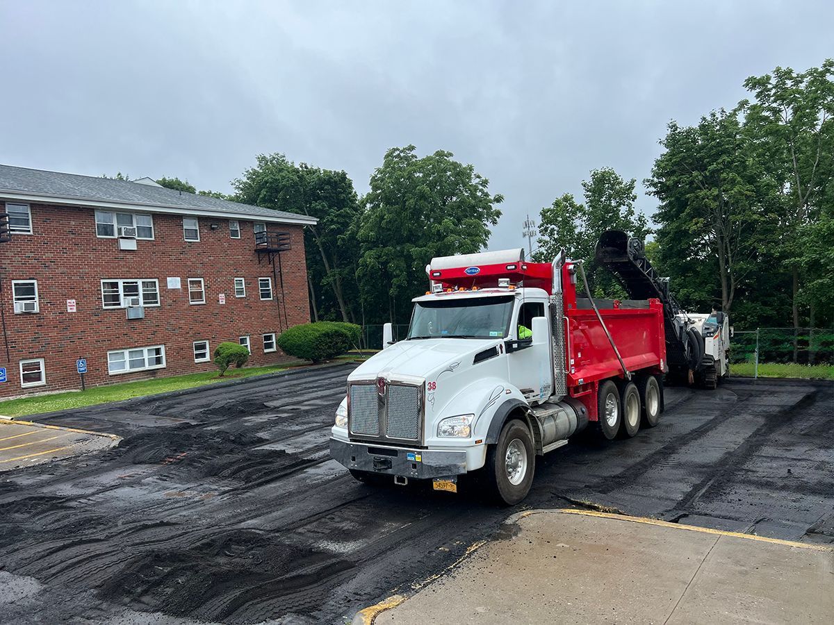Asphalt paving in progress: red dump truck and paving machine on a parking lot next to an apartment building.