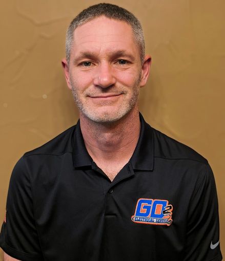 Portrait of a man in a black polo shirt with a blue-orange logo against a tan background
