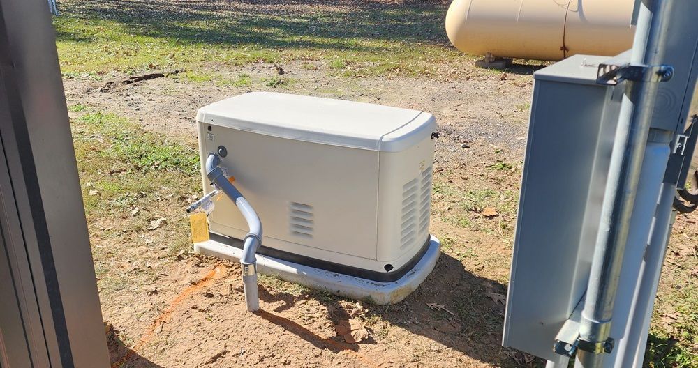 White outdoor generator beside a metal pole on dirt and grass near a tank and fence.