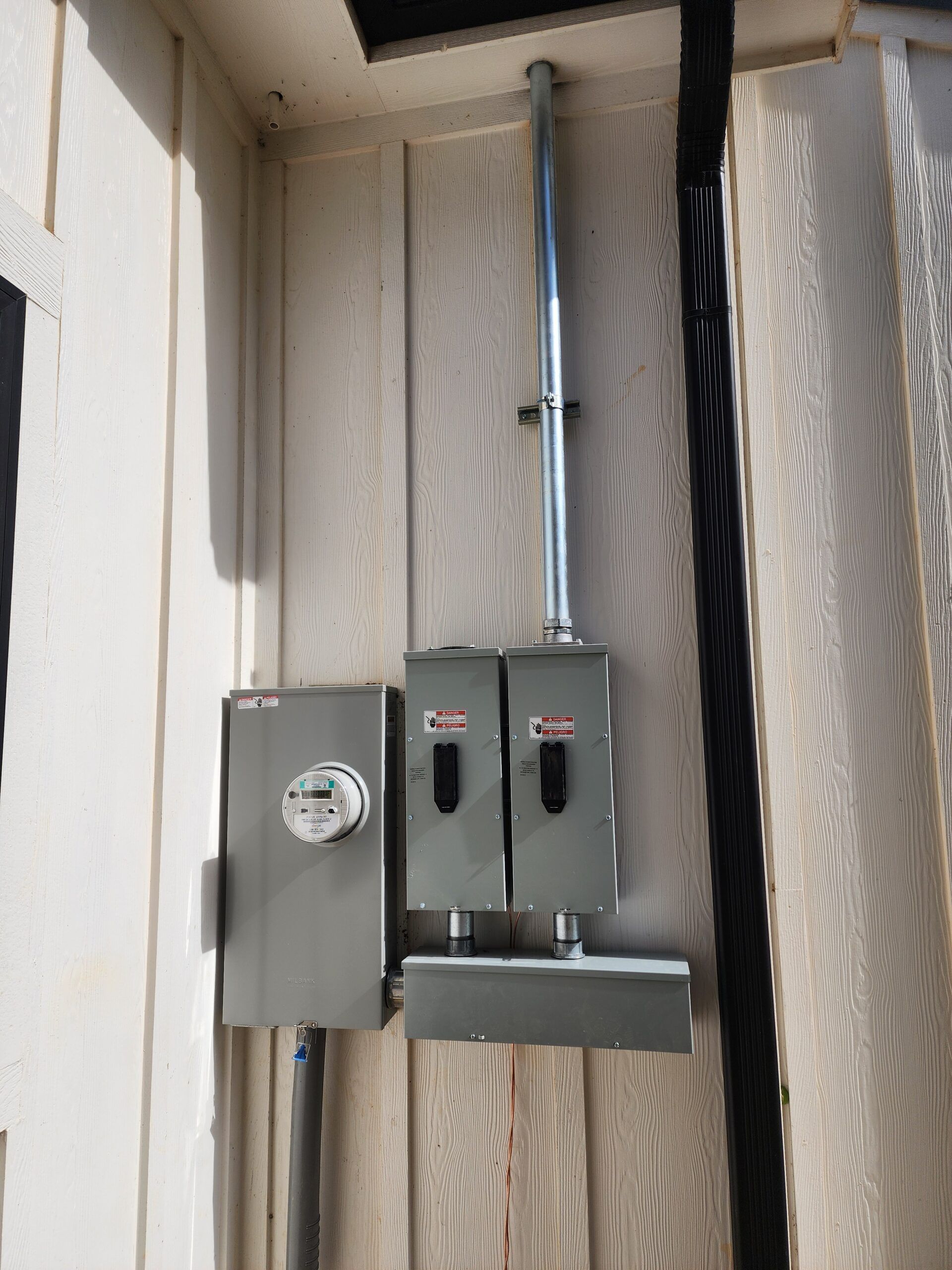 Gray electrical meter boxes and conduit mounted on an exterior wall next to a wooden fence.