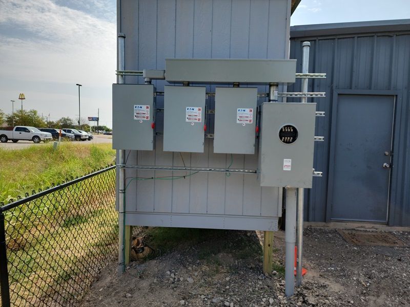 Three gray electrical utility panels mounted on a metal stand beside a blue building and chain-link fence.