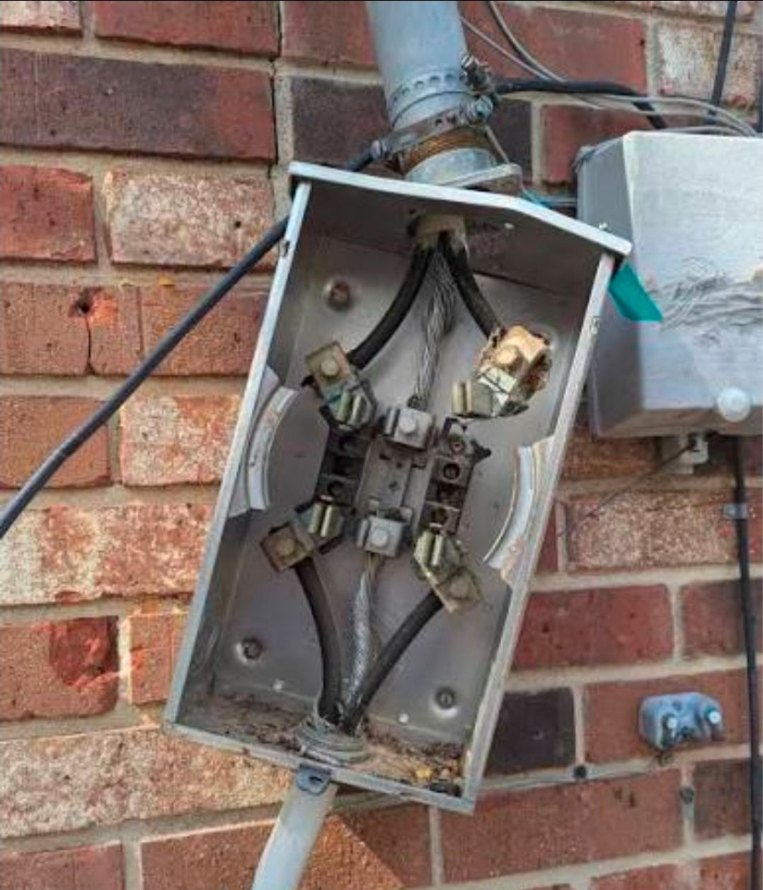 Damaged electrical meter box on a brick wall with exposed wiring and broken cover