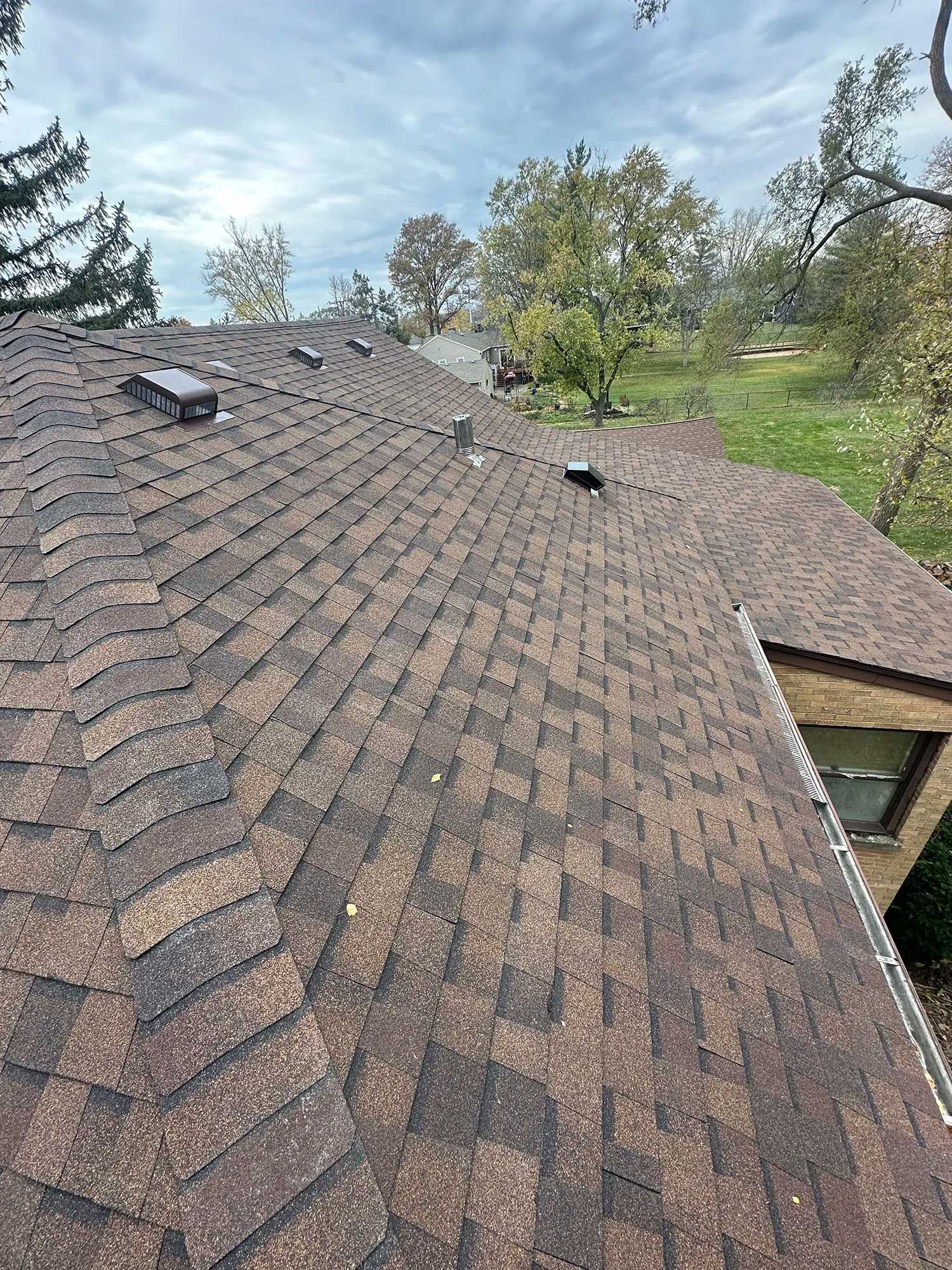 Brown asphalt shingle roof with vents, set against a cloudy sky, trees and a brick building.