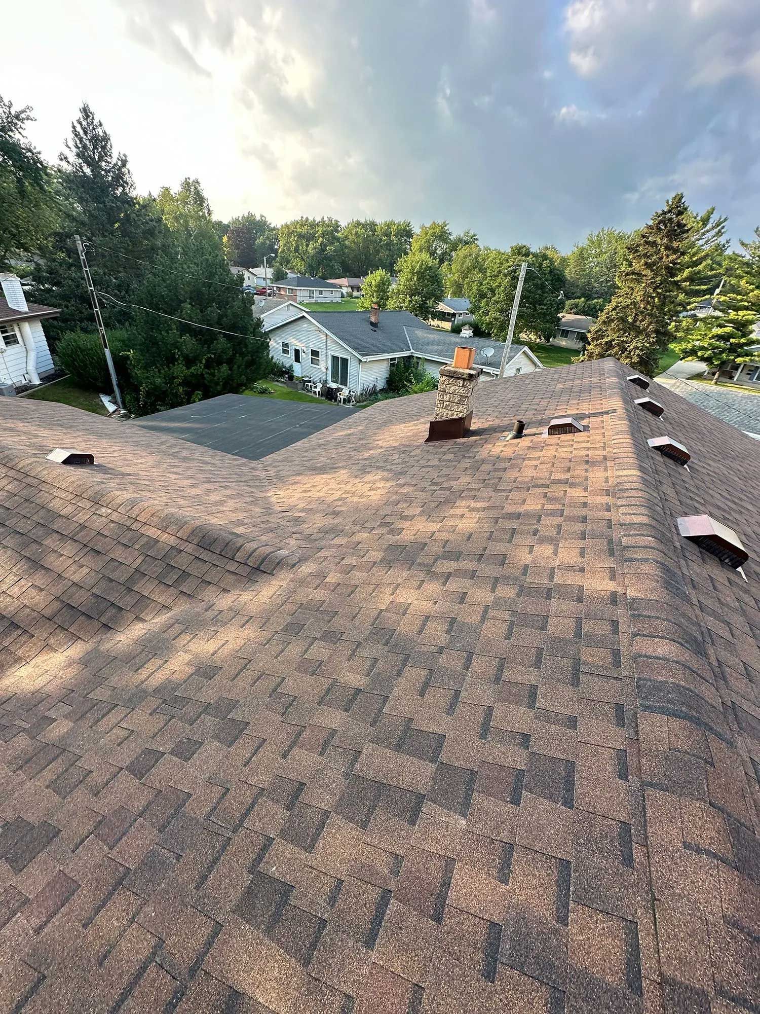 Brown shingled roof, with vents and chimney, overlooking houses and trees under a cloudy sky.