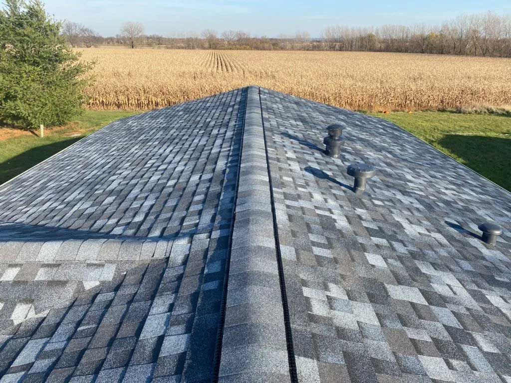 View of a new roof with gray shingles, a central ridge, and vents, with a field in the background.