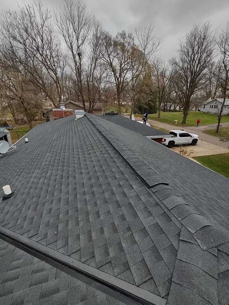 Gray asphalt shingle roof of a house with a dark metal ridge cap, overcast day.