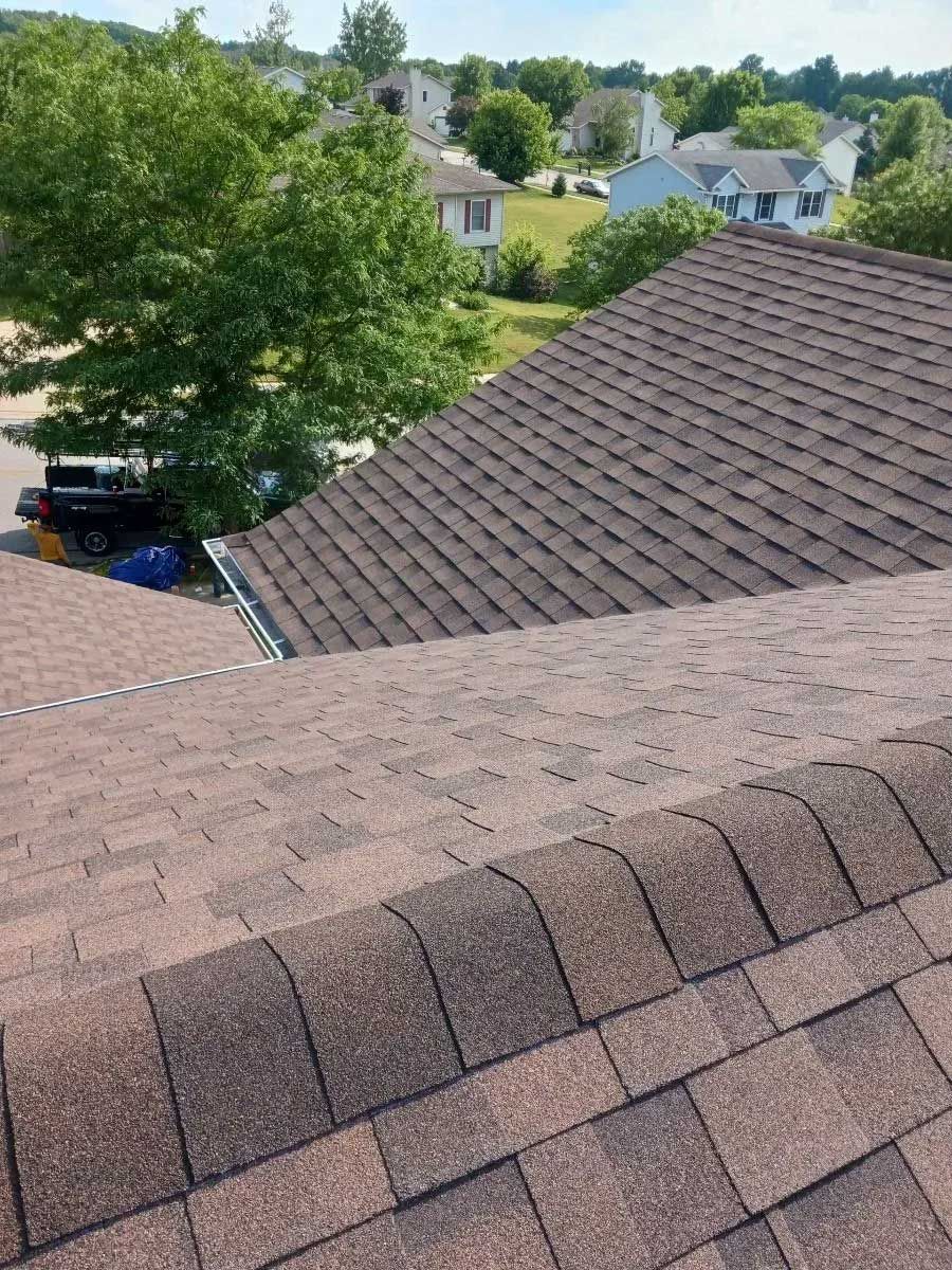 Brown shingle roof with valley, treetops, houses in the distance under blue sky.