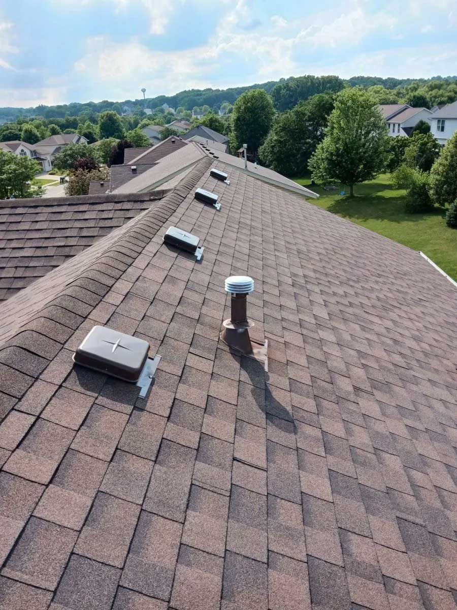 Brown shingled roof with vents, under a blue sky, overlooking a residential area.