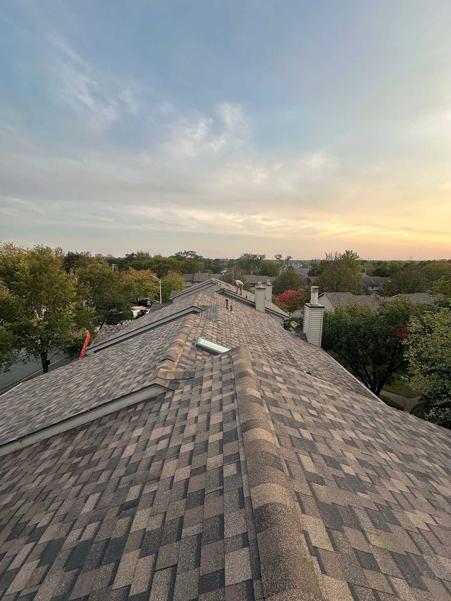 Rooftop view of a neighborhood at dusk; brown shingle roof with a blue and yellow sky.