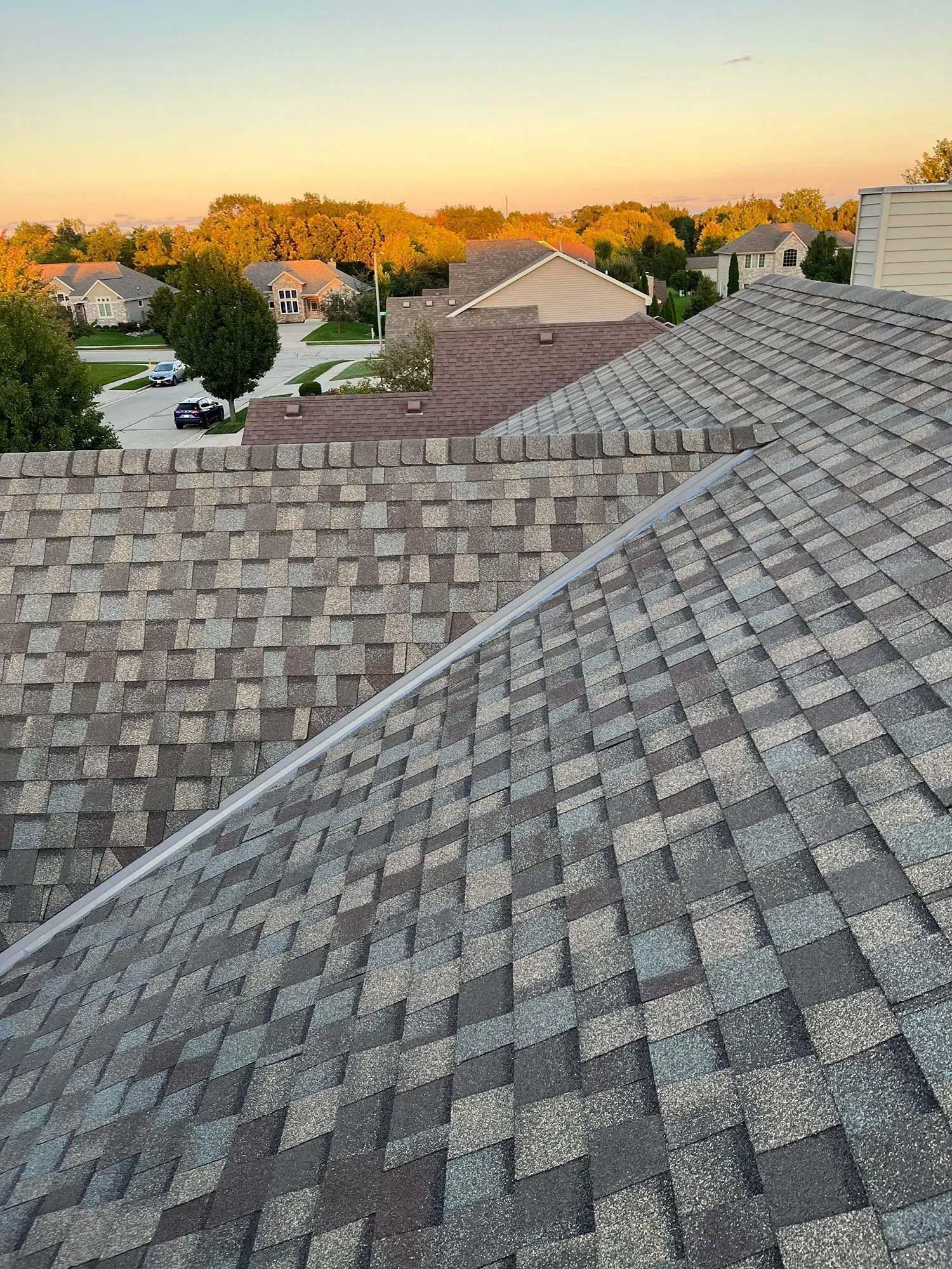 View of rooftops in a suburban area at sunset, with focus on the gray-shingled roof in the foreground.
