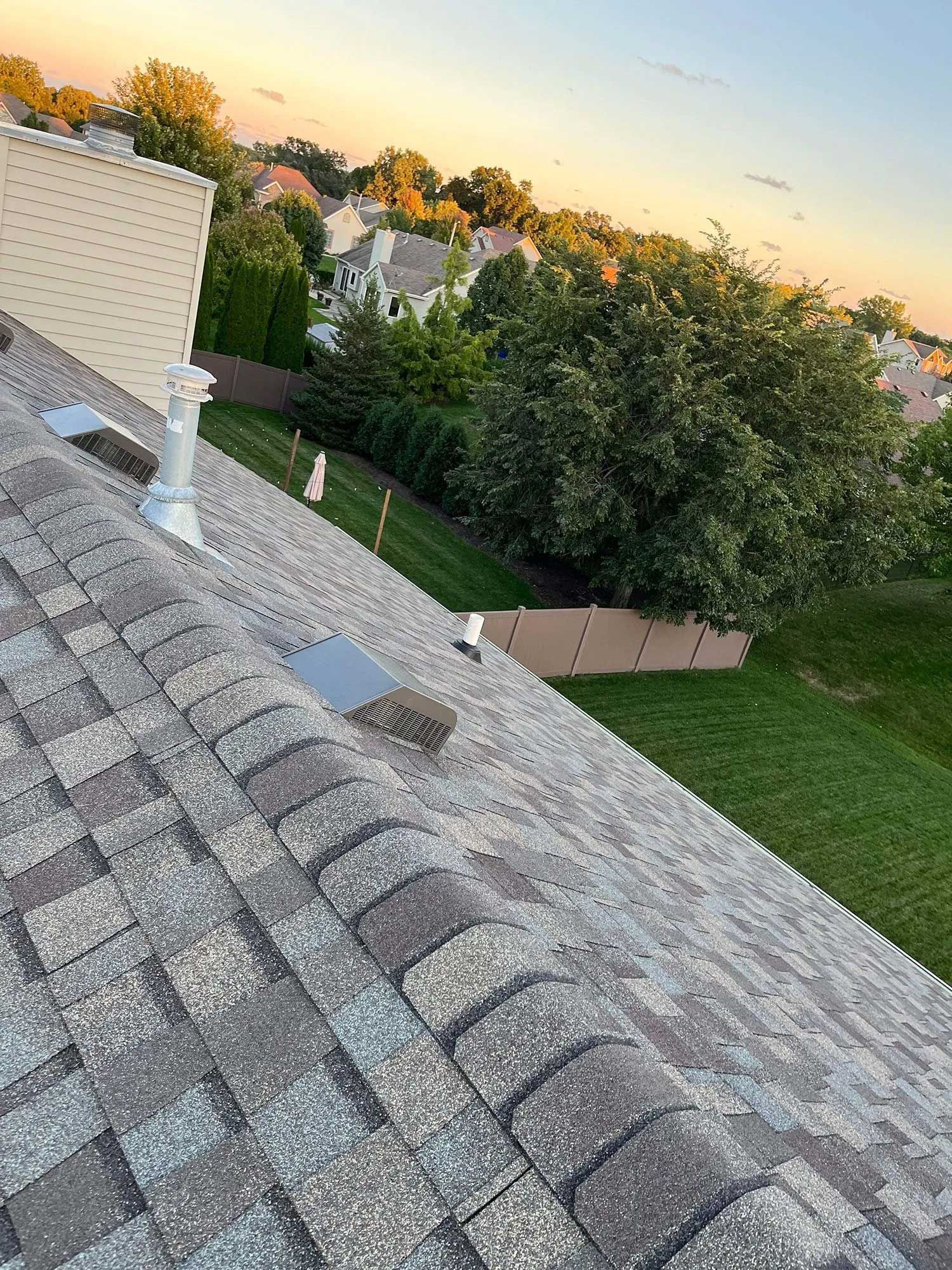 A roof with brown shingles slopes toward a green lawn and trees at sunset.