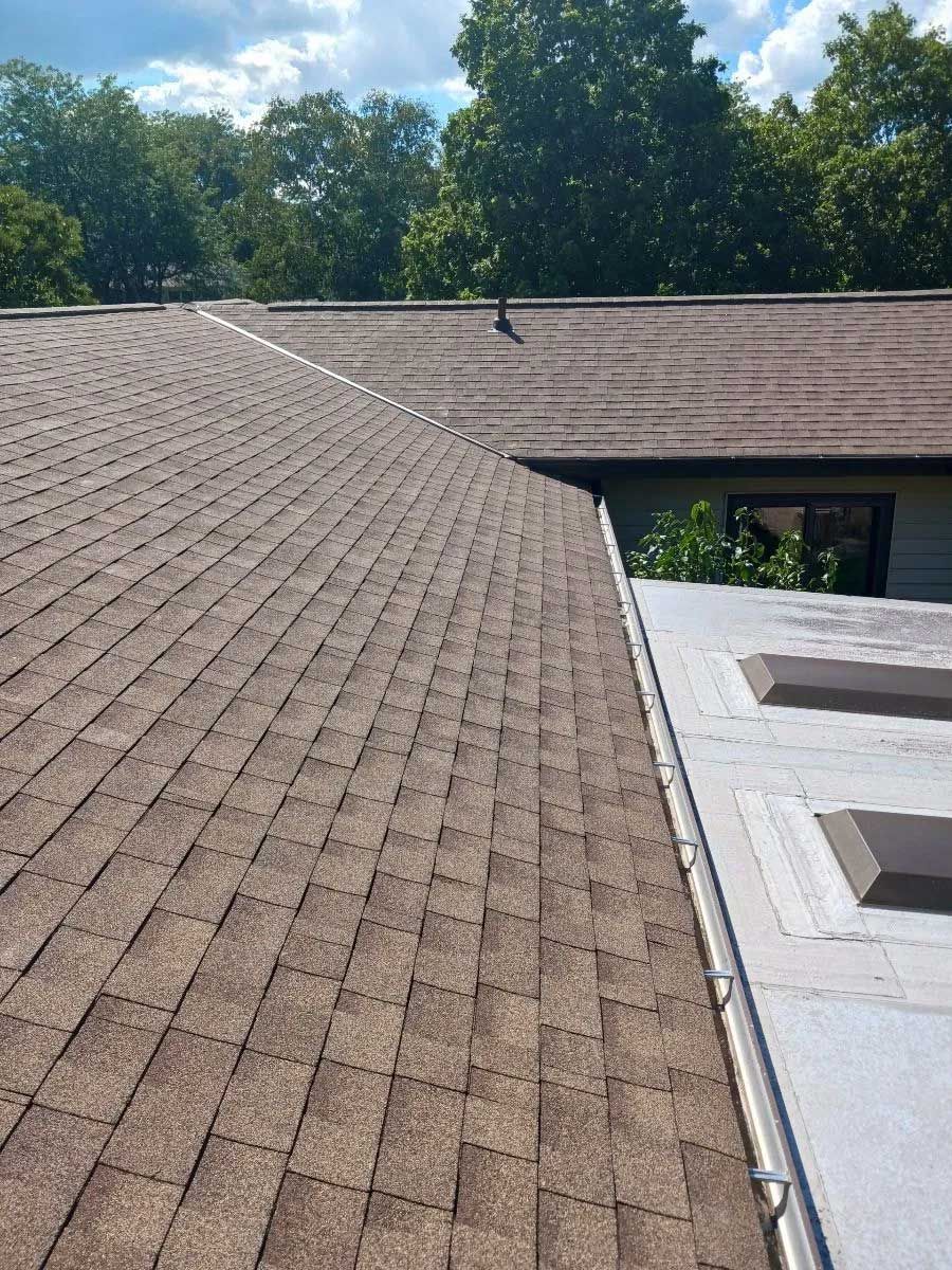 Brown shingle roof meets a flat roof, trees in the background under a cloudy sky.