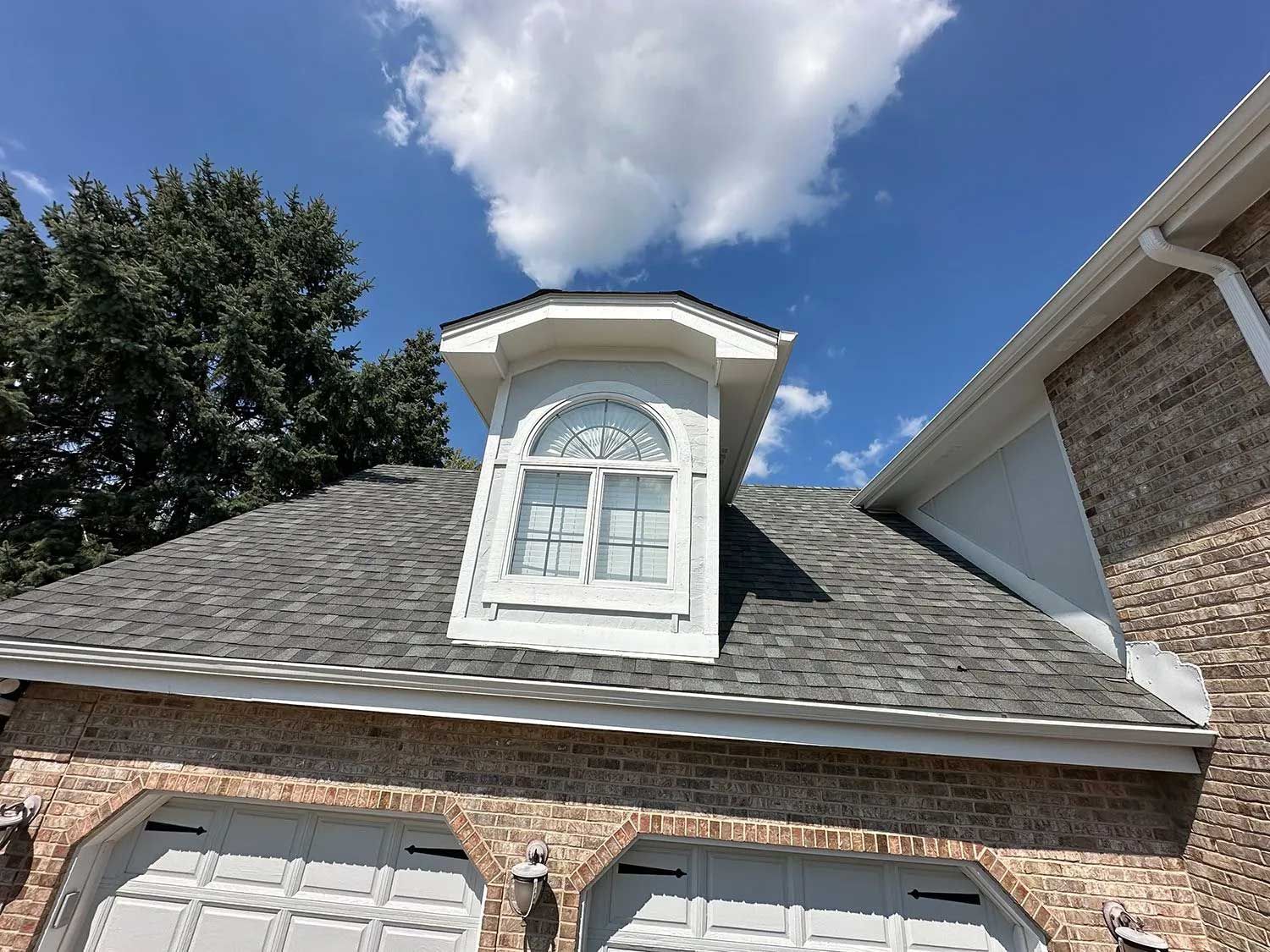 Dormer window on a brick house with a gray shingle roof against a blue sky with clouds.