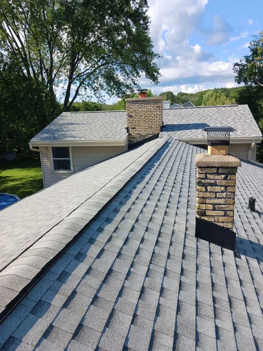 Gray shingled roof with two brick chimneys, trees and blue sky in the background.