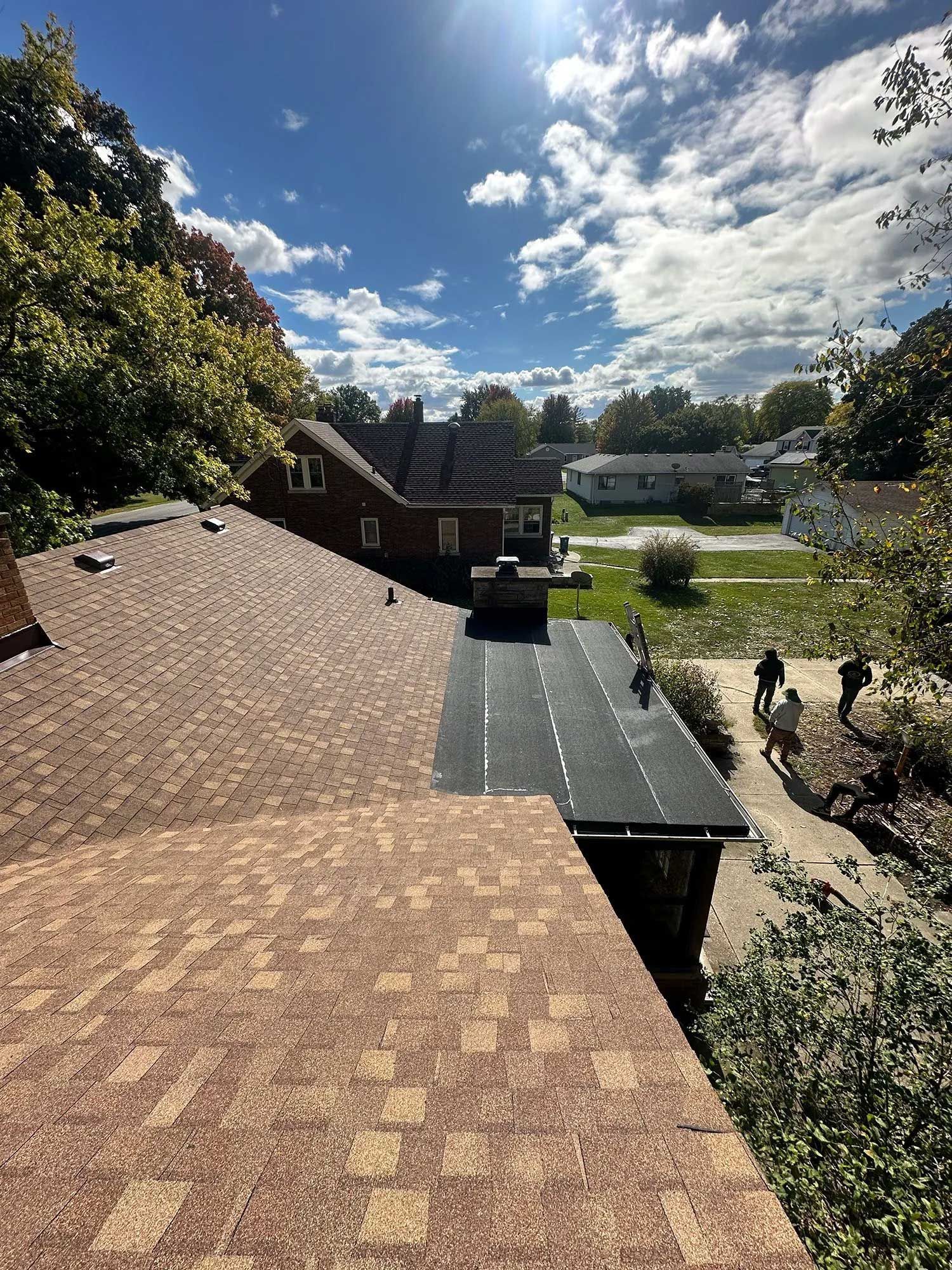 View of a brown shingle roof in progress, blue sky, and two people working in the background.