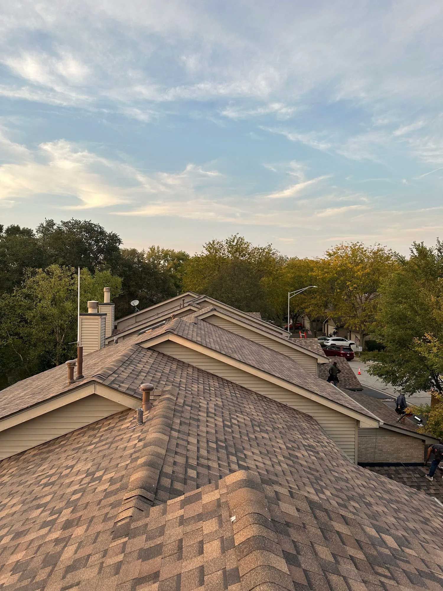 View of rooftops with brown shingles, chimneys, and trees against a cloudy blue sky.