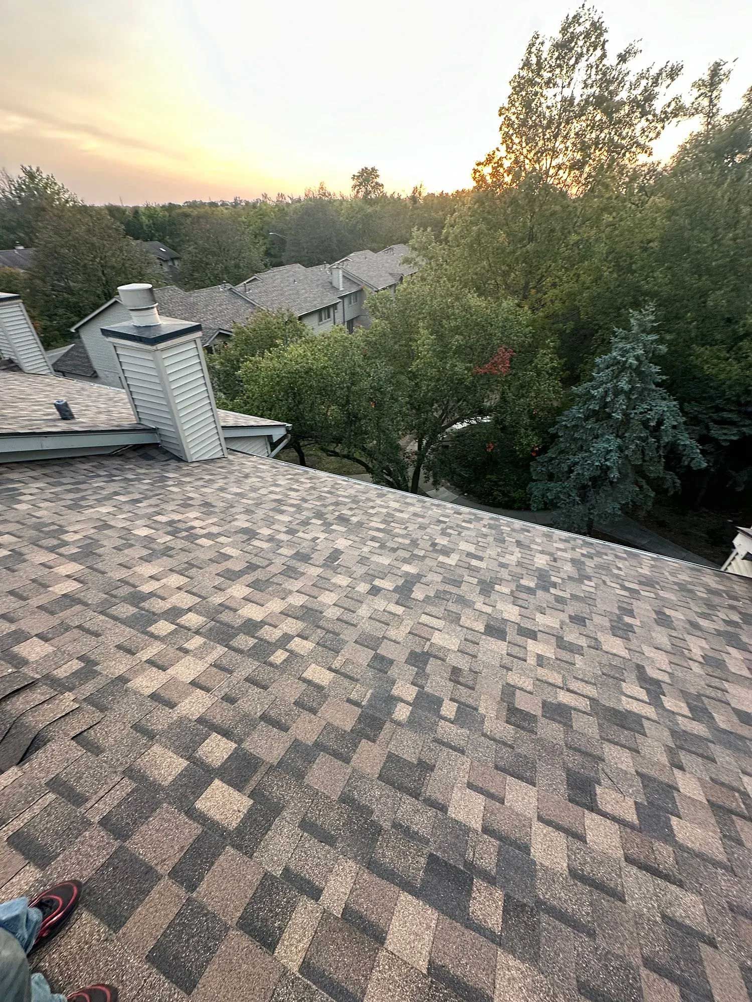 Rooftop view with brown and grey shingles, chimney, trees, and houses in background; sunset.