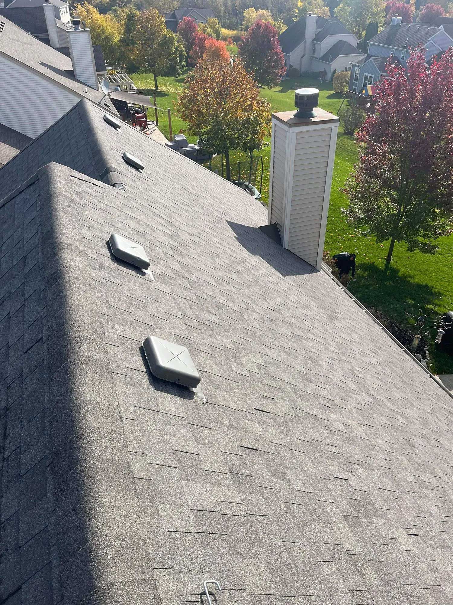 A gray asphalt shingle roof with multiple vent stacks. Green grass, trees, and houses are visible in the background.