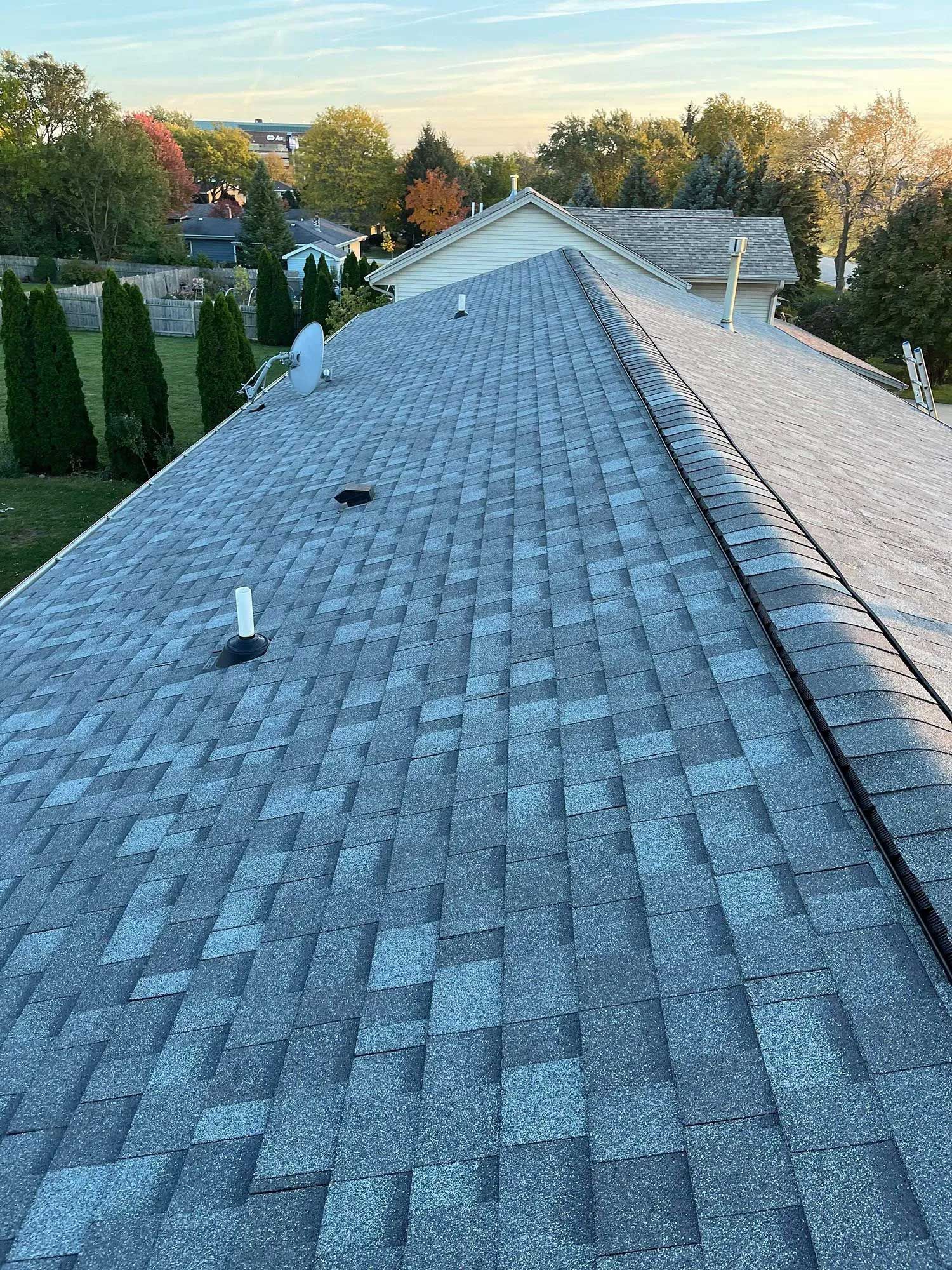 Gray shingled roof with a variety of vent pipes, set against trees and sky.