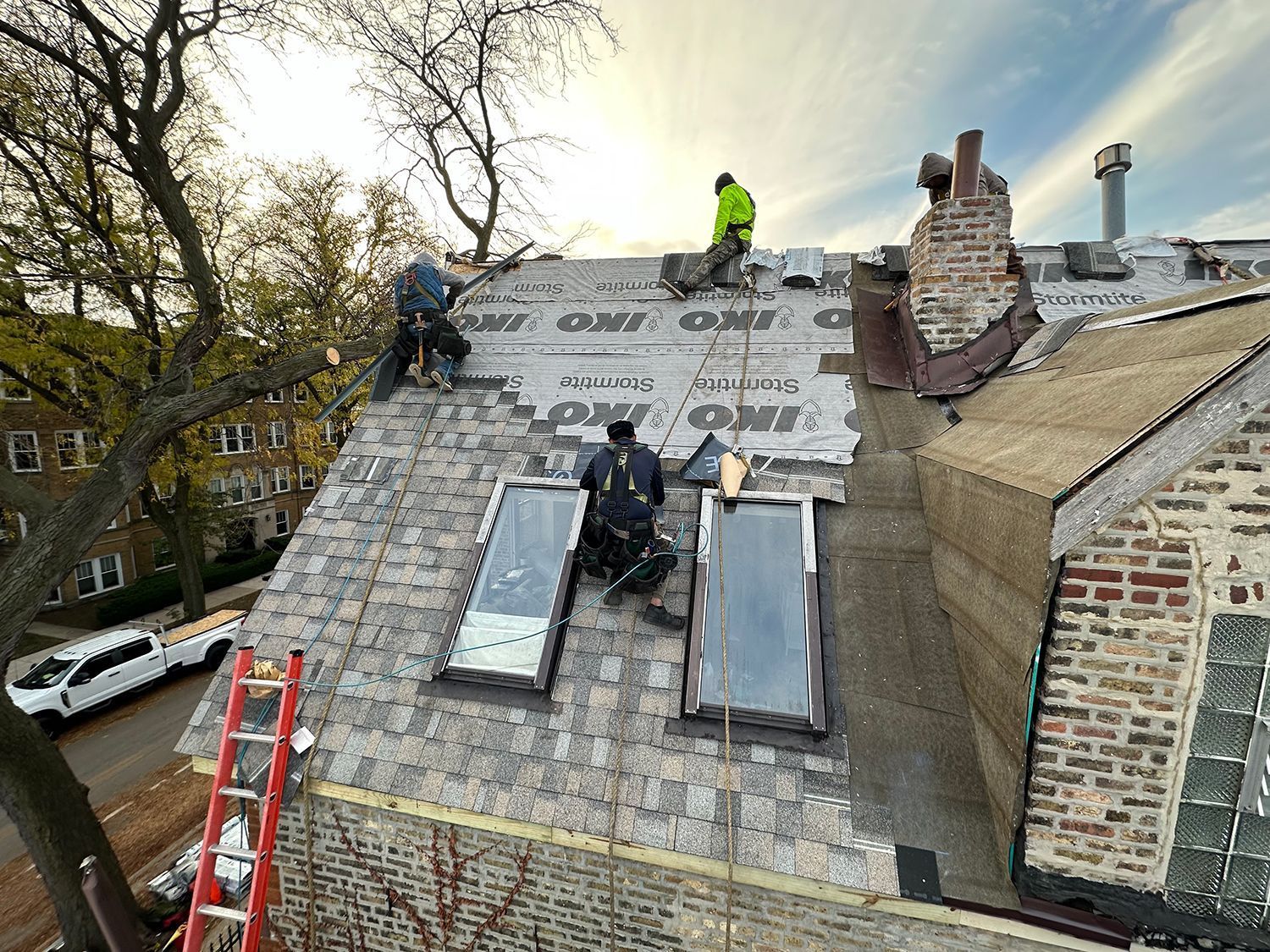 Roofers working on a residential roof, installing new shingles. They are on a cloudy day, near a chimney.