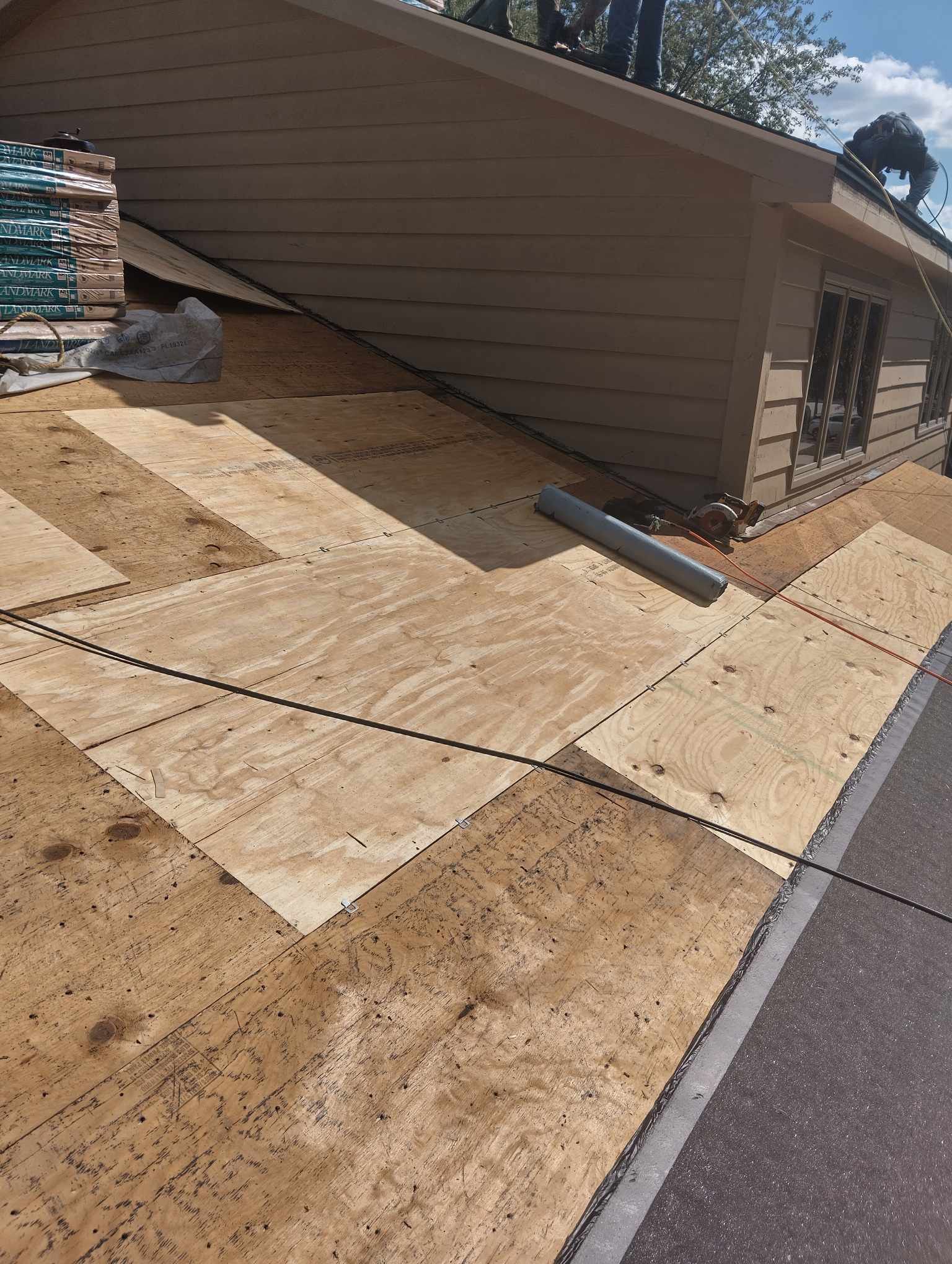 Roof partially covered with plywood, workers visible. Brown roof, tan siding, materials present.