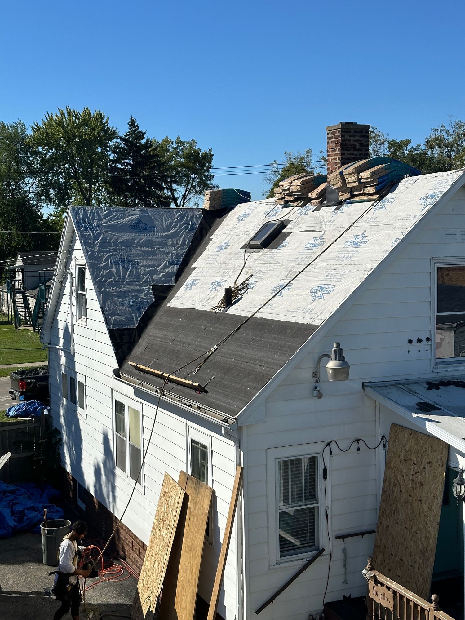 House roof under construction; shingles removed, tar paper visible; brick chimney, blue sky.