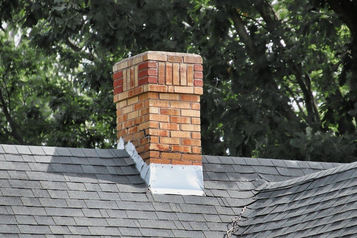Brick chimney on a gray shingle roof with trees in the background.