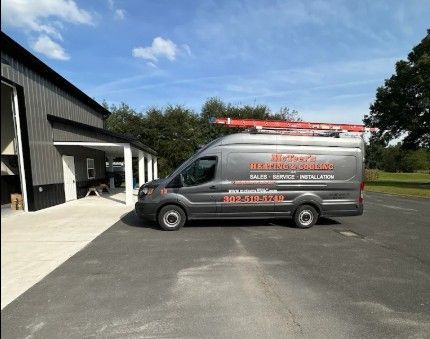 A grey work van with business branding parked on an asphalt lot beside a modern metal warehouse building.