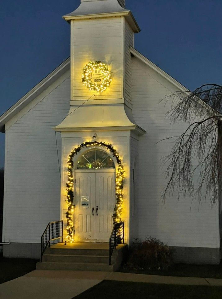 A white church entrance at dusk, decorated with a glowing wreath above the door and illuminated garland around the frame.