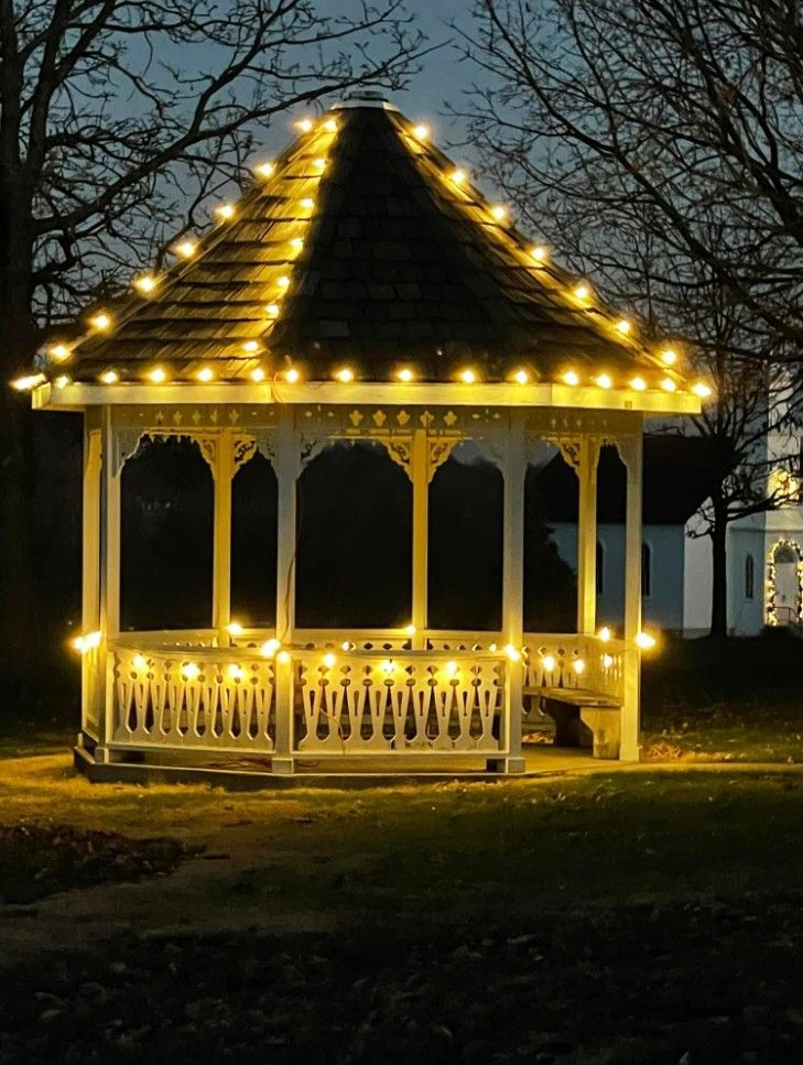A white gazebo at night, outlined with glowing warm string lights in a park setting.