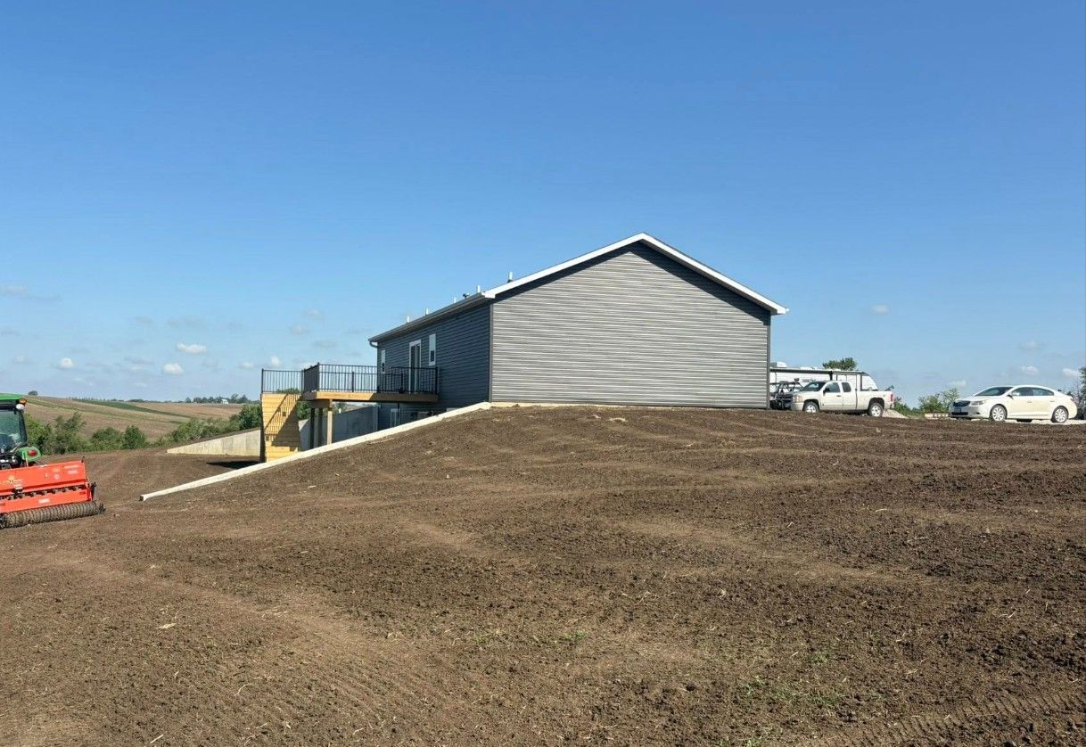 A newly constructed gray building sits atop a graded hill of dark soil with vehicles parked nearby.