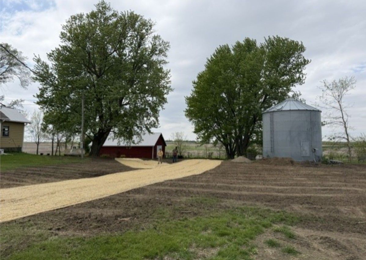 Rural farmyard with gravel drive, two leafy trees, a red shed, and a metal grain bin under cloudy sky