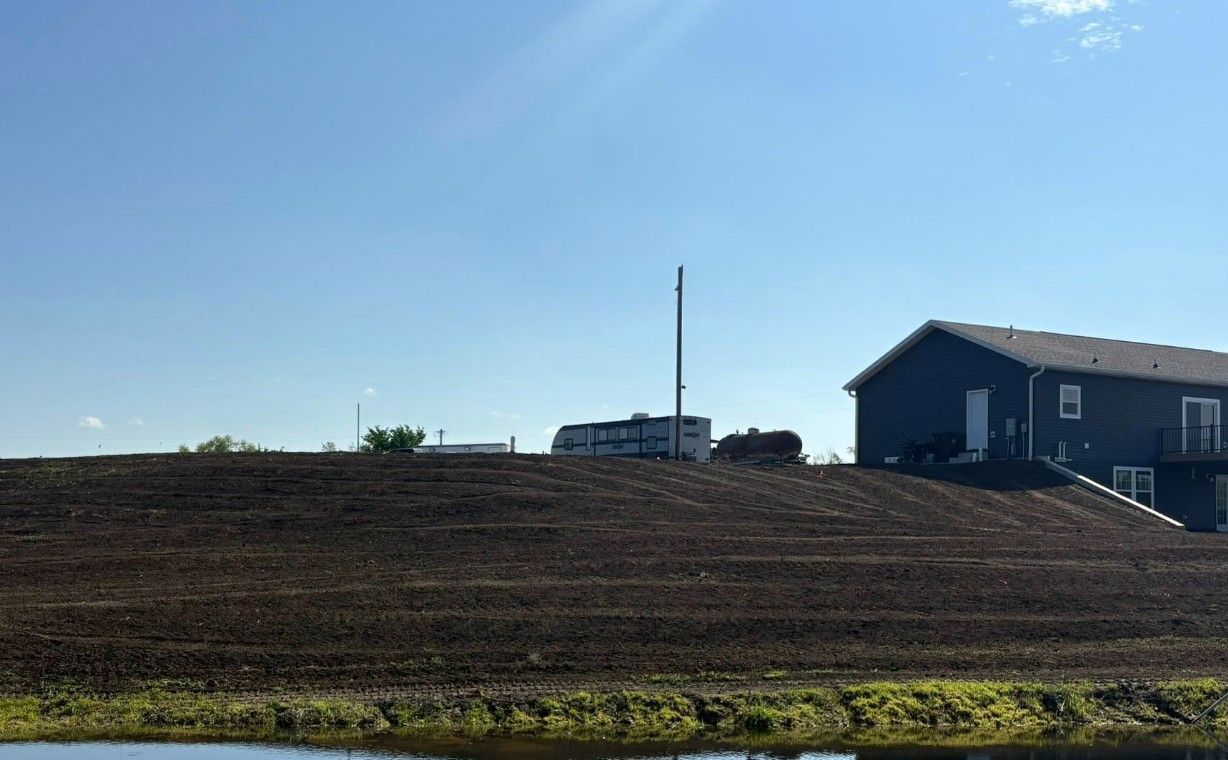 Earthen berm with a house and pond under a blue sky with some wispy clouds.