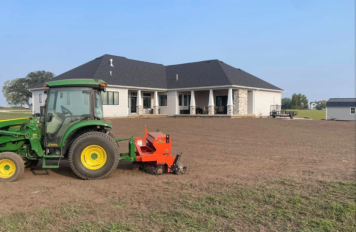 Green tractor with tiller, working on a prepared field in front of a house.