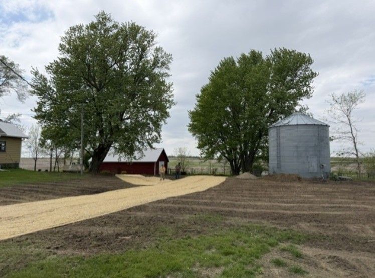 Gravel farm lane between trees and a red shed, leading past a metal silo in a cloudy rural field