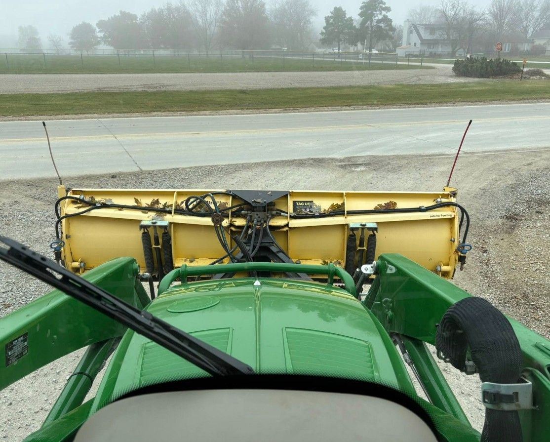 Green tractor with a yellow snowplow on a gravel road, foggy background.