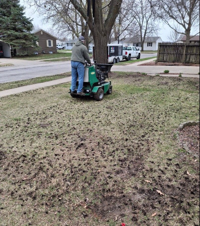 Man on aerator machine, working on lawn with dark patches, street and houses in background.