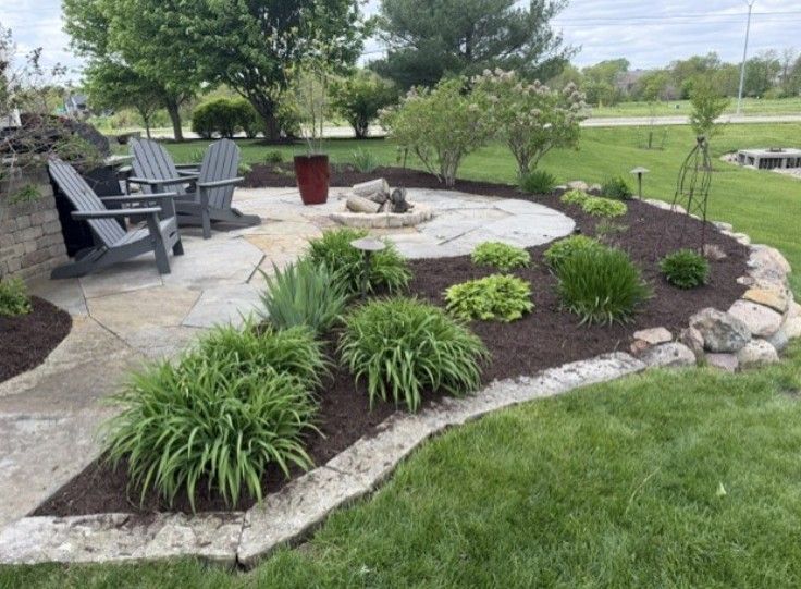 Patio seating beside a stone fire pit and landscaped garden with mulch, shrubs, and green lawn.