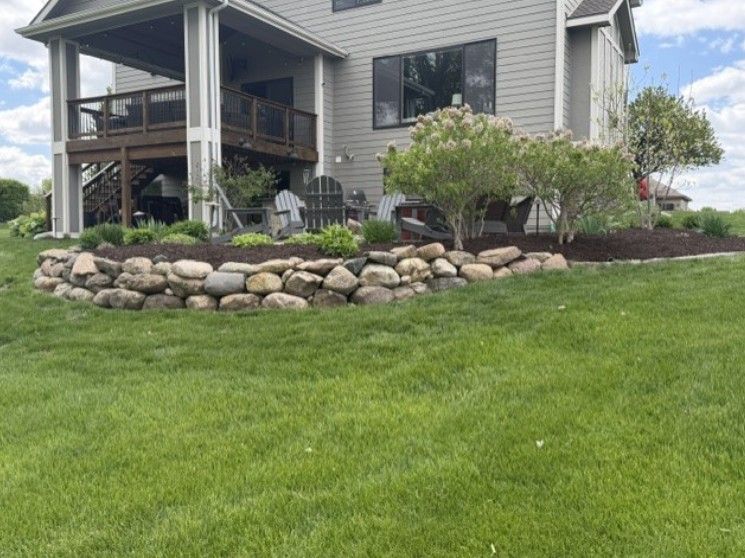 Gray house with covered porch, landscaped shrubs, and a stone border above a green lawn.