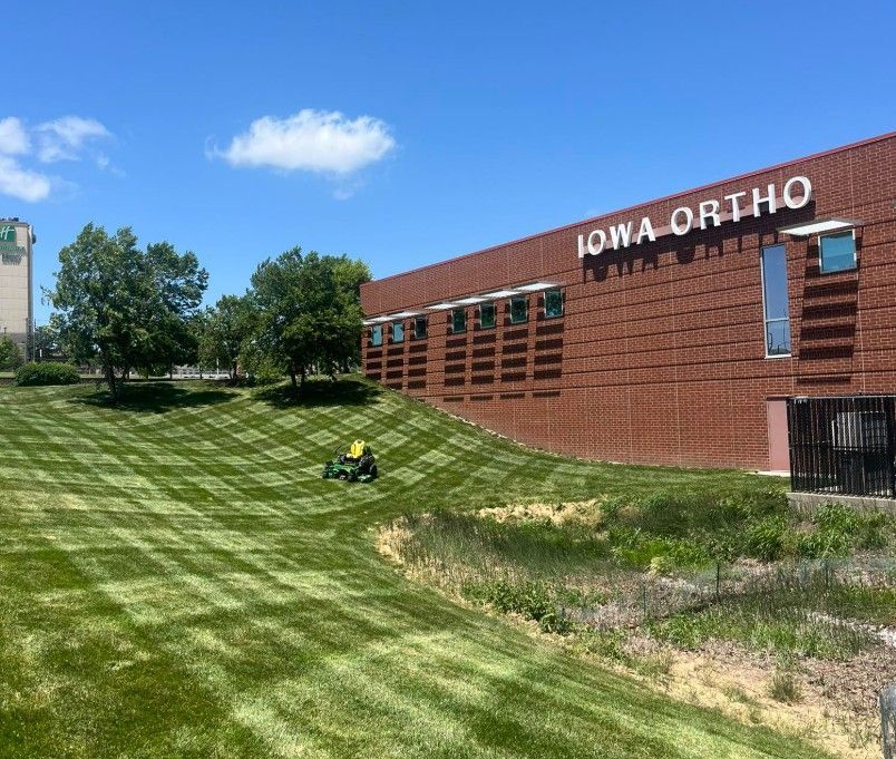 Lawn mower cuts grass on a sloped yard by the Iowa Ortho building under a blue sky.
