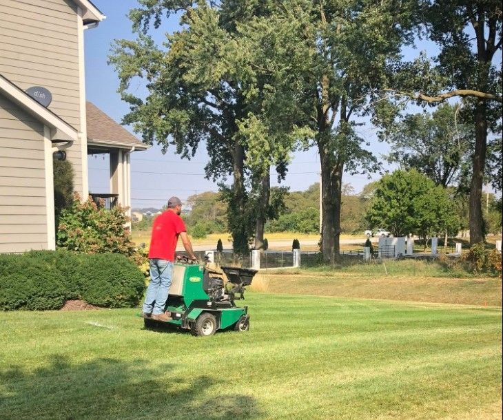 Man operating a green lawn spreader on a freshly mowed lawn next to a house.