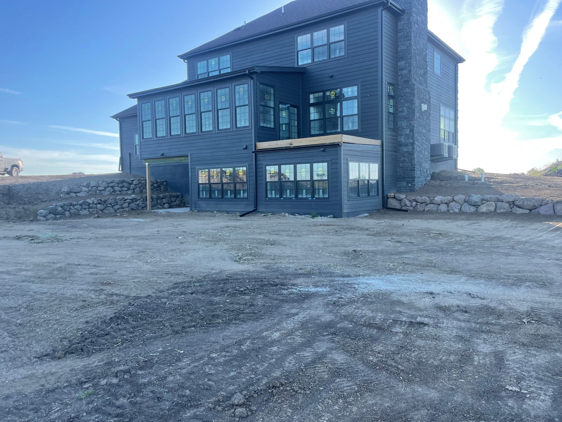 Modern dark stone house with large windows on a barren, rocky lot under a bright blue sky