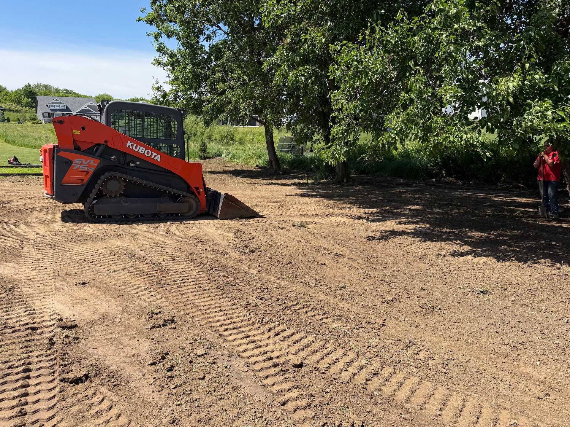 Orange skid steer on dirt lot near trees, with a person standing at right.