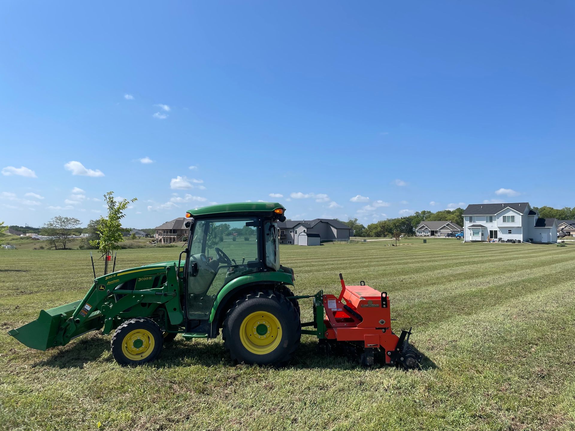 Green tractor pulling red mower across a grassy field under a blue sky