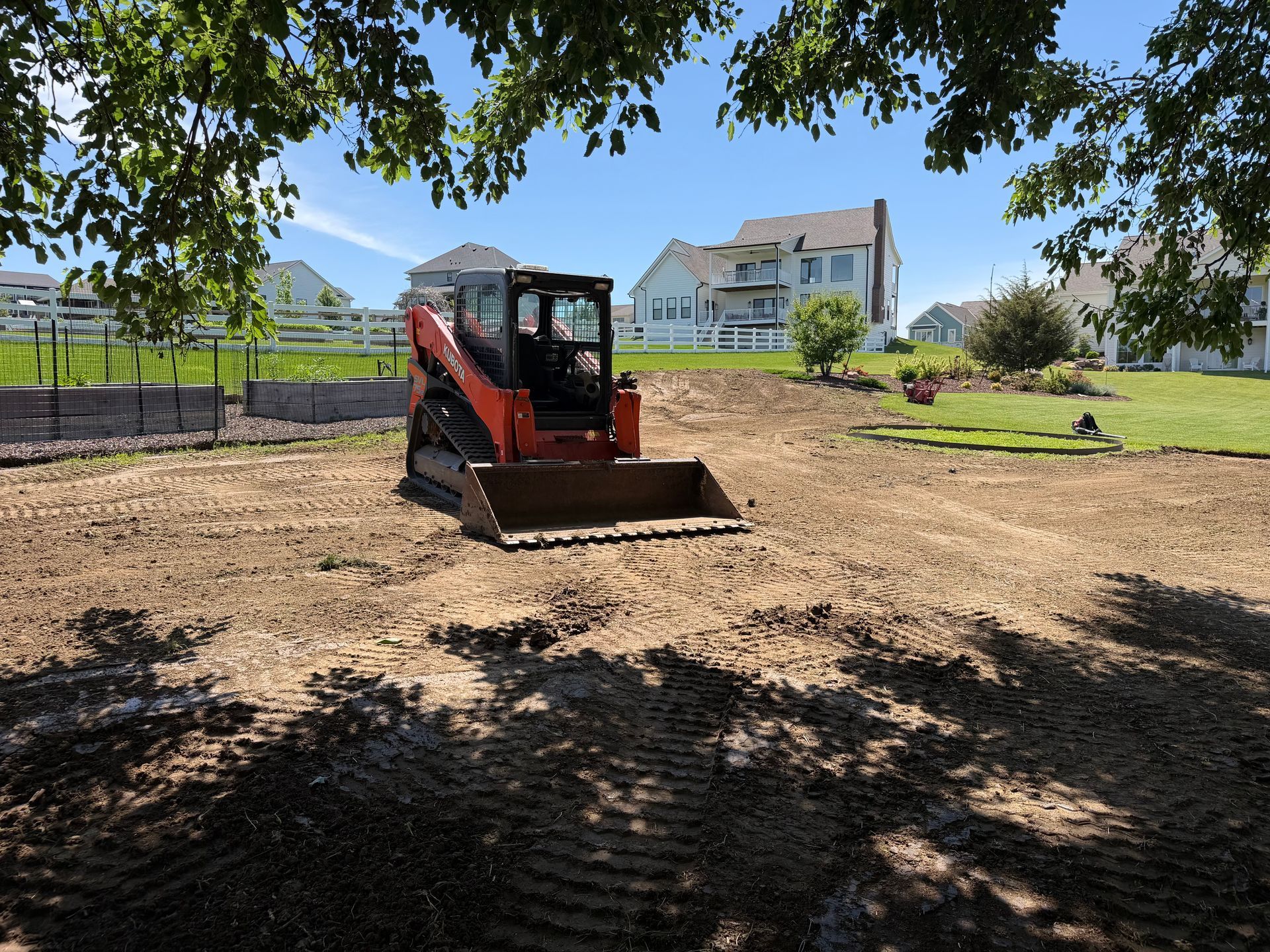 Orange skid steer on dirt lot near houses, with trees and mountains in the background
