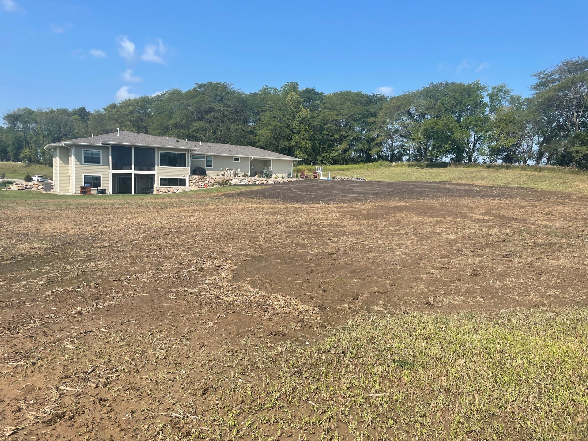 Bare dirt lot in front of a small white building under a blue sky, with trees in the background