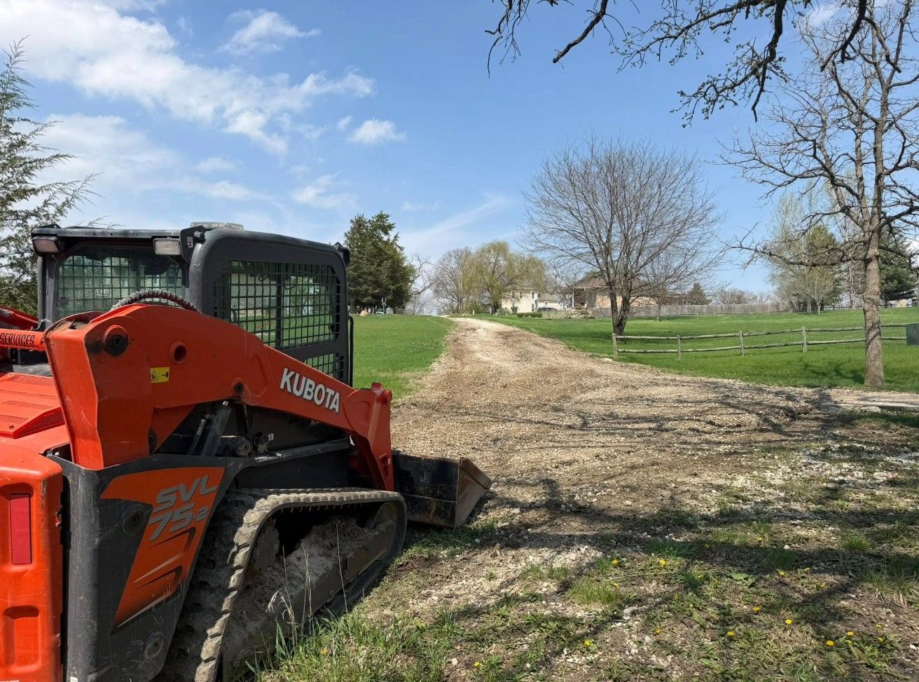 Orange Kubota track loader clearing dirt on a grassy path under a blue sky.