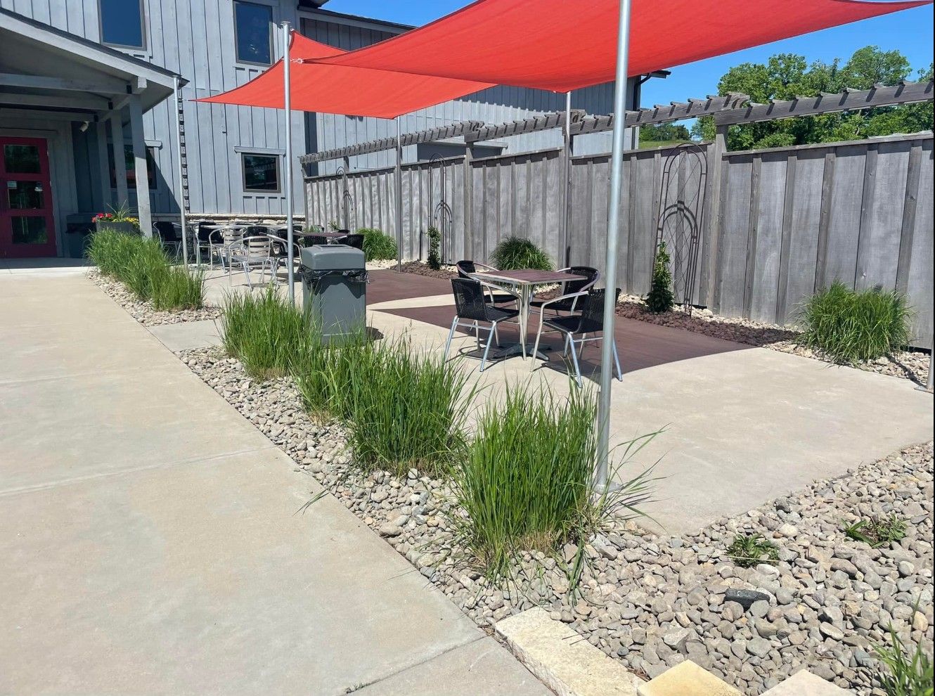 Outdoor patio with red shade sails, a concrete walkway, gravel landscaping, and table seating next to a wooden fence.