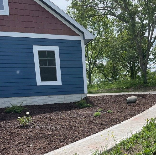 Blue house side with white-trimmed window, mulch bed, and curved concrete walkway under trees